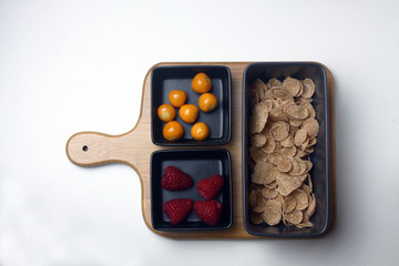 raspberry, cape gooseberry and cornflakes in black bowls on wooden cutting board on white background flat lay