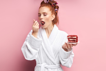Attractive woman wearing bathrobe, curlers after shower eating sweet tasty cake. Emotional girl posing with cake isolated over pink background