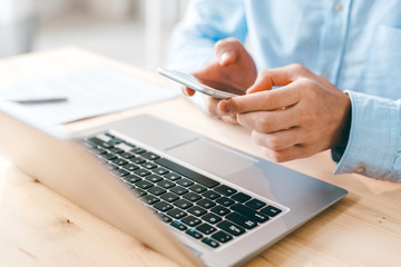 Young male employee scrolling or texting in smartphone over laptop keypad