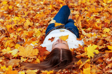 Adult woman feels hassle-free like a little girl concept. Lady in blue jeans and white sweater lies on fallen yellow dry autumn leaves. Head on the foreground. Rest in the park and forest.