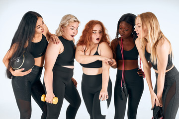 Five attractive girls stand together isolated over white background, smile and talk. Redhaired girl show ring, other girls look and surprised. Different nations, nationalities