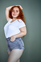 Cute girl with curly natural red hair stand posing isolated over grey background. Portrait, wearing denim shorts, white t-shirt. Natural beauty