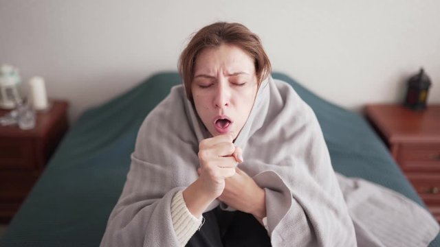 An unhealthy young woman with a sore throat and fever takes cover with a blanket in bed in the bedroom. Heating problems during the flu season. Sick leave, cold symptoms