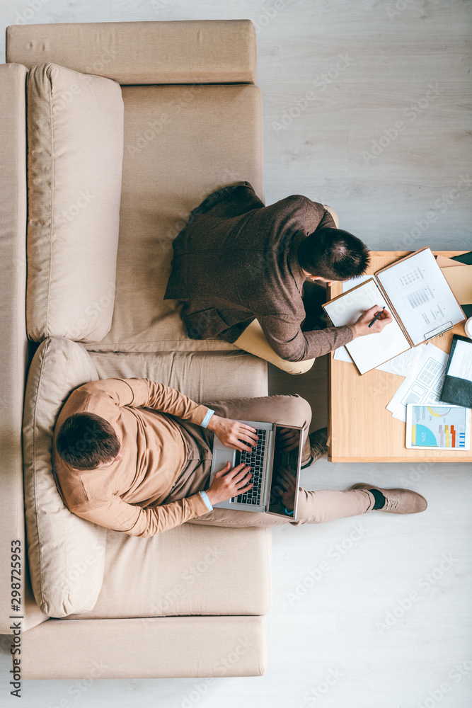 Poster Young broker making notes while his colleague with laptop working over project