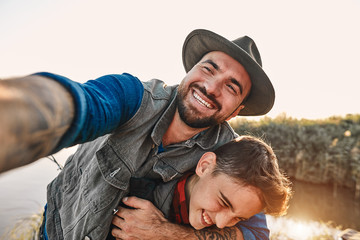 Father hugs son and takes selfie. They smile broadly and enjoy. Background lake.