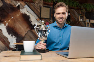 Joyful businessman holding his trophy and working on his laptop
