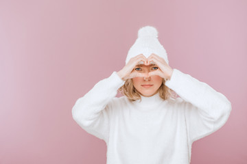 Pretty girl looking through heart gesture made with hands while standing isolated over pink background. The girl is dressed in a fluffy sweater and a knitted hat.