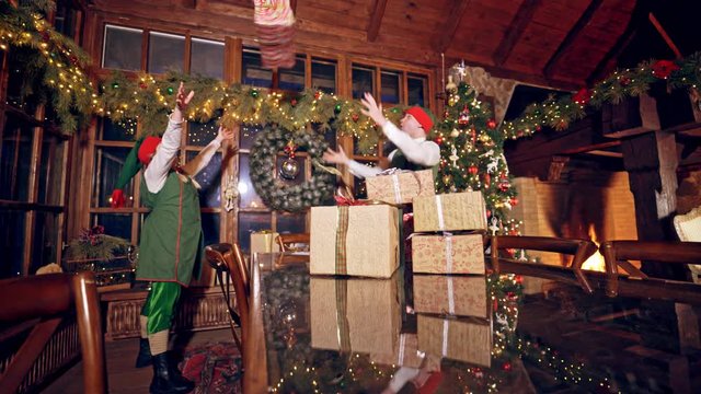 Joyful elves with gift boxes. Happy fairy characters making a pyramid of presents on the table on Christmas decorations background.