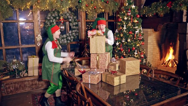Happy elves with many presents indoors. Christmas decorated room and elves in green costumes putting packed gifts on the table. Christmas time.
