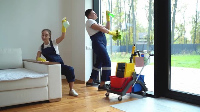 Cheerful young woman nd man from cleaning service dressed in workwear and rubber gloves dancing in house