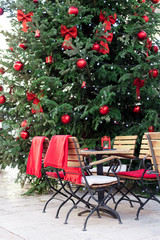 Street cafe under Christmas tree with red ornaments outdoor. Wooden tables and chairs with warm plaid at winter market in old european town. Cozy festive and New Year atmosphere in Europe