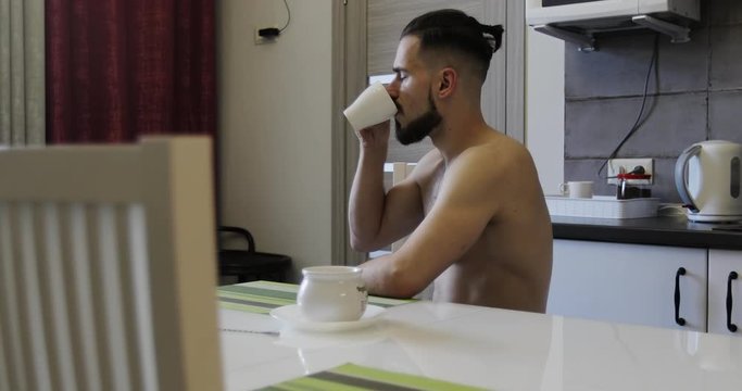 A bearded man is drinking coffee sitting at a table in the kitchen.