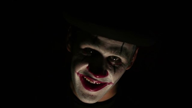 Terrible clown looks at the camera and laughs terribly. A terrible man in a clown makeup waves his hand and beckons his victim with a finger, looks at the camera and laughs. Scary clown grimaces looki