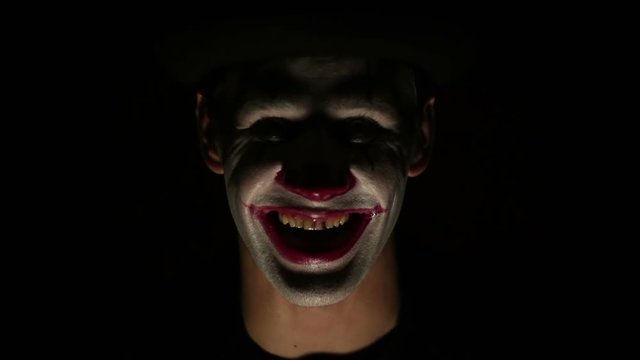 Terrible clown looks at the camera and laughs terribly. Scary grimaces of a clown looking at the camera. Halloween Terrible man in the makeup of a clown raises his head and opens his eyes, looks at th