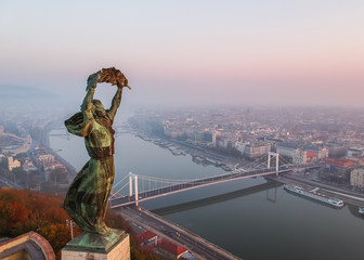 Naklejka premium Aerial view to the Statue of Liberty with Elisabeth Bridge and River Danube taken from Gellert Hill on sunrise in fog in Budapest, Hungary.