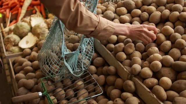 Close-up female hands picking potatoes in a string bag from wooden boxes in a greengrocery. Eco packaging, zero waste