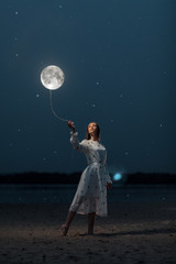 Young attractive girl in a long white dress holds the moon on a rope. Art photography