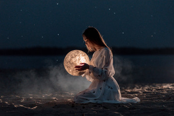 Beautiful young girl on a night beach with sand and stars holds the moon in her hands