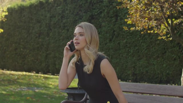 A beautiful girl of European appearance speaks on the phone in the park. A woman first sits on a bench, and then goes along the alley (shooting from the back)