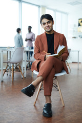 Full length portrait of young Middle-Eastern businessman smiling at camera while sitting on chair in modern office
