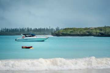 Boats on the water and waves crashing on the beach on a slightly overcast morning at Kuto Bay on Isle of Pines in New Caledonia- French Polynesia, South Pacific.