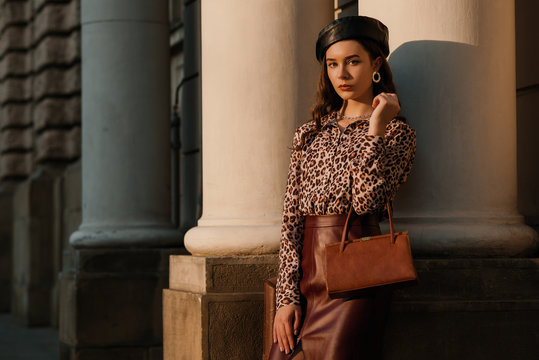 Outdoor fashion portrait of young elegant luxury woman wearing leopard print shirt, faux leather skirt, beret, holding small brown classic bag, handbag, posing in street Copy, empty space for text 