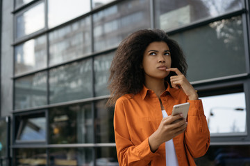 Pensive woman holding smartphone, shopping online, ordering food, booking tickets, searching information on website.  Young beautiful African American girl using mobile phone, waiting for taxi  