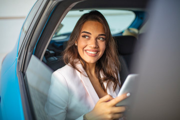 Young, beautiful woman sitting in the back seat of a car with a tablet in hands. Beautiful woman is using a tablet in car. She is sitting on back seat and relaxing.