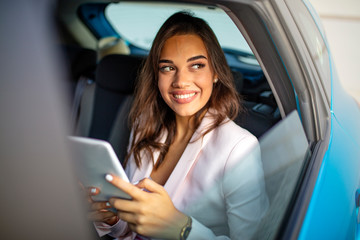 Fototapeta na wymiar Young, beautiful woman sitting in the back seat of a car with a tablet in hands. Beautiful woman is using a tablet in car. She is sitting on back seat and relaxing.