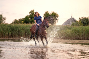 Fototapeta premium A young girl riding a horse on a shallow lake. A horse runs on water at sunset. Care and walk with the horse. Strength and Beauty