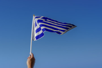 Hand waving Greek flag in the air for a national celebration. Blue & white Greek plastic flag with...
