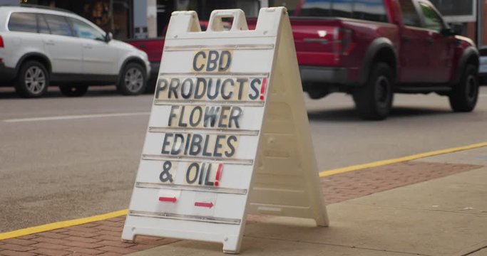 A daytime view of a sign outside of a CBD products store on the Main Street of a small town. Traffic passes in the background.  	