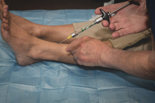 Podiatrist Or Foot Surgeon Performs An Ankle Injection Of Local Anaesthetic To Numb The Food For Awake Surgery Or Procedure. 