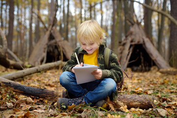 Little boy scout is orienteering in forest. Child is sitting on fallen tree and writing in the...