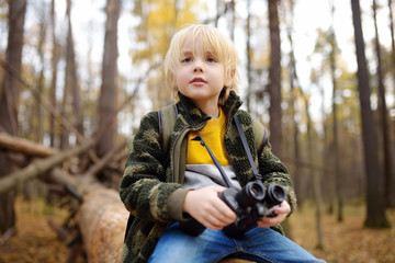 Little boy scout with binoculars during hiking in autumn forest. Child is sitting on large fallen tree and looking through a binoculars.