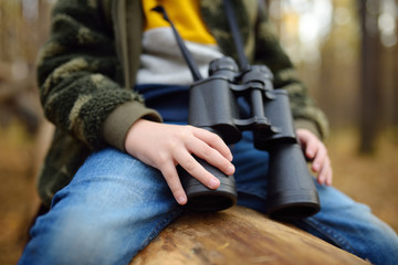 Little boy scout with binoculars during hiking in autumn forest. Child is sitting on large fallen tree and looking through a binoculars.