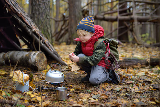 Little boy scout during hike in forest on autumn day. Child is cooking tea with help tourist gas burner. Behind the child is teepee hut.