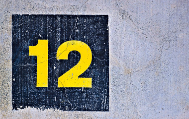 Yellow number 12, twelve, in dark blue square, offset on flat concrete surface.