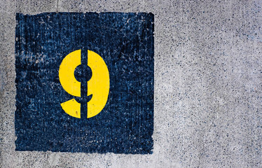 Yellow number 9, nine, in dark blue square, offset on flat concrete surface.
