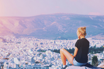 Fototapeta na wymiar Young tourist woman sitting on top of mountain and looking at a beautiful landscape cityscape Athens Greece. Adult girl tourist relax on hill overlooking Athens in summer. Famous Athens city in Europe