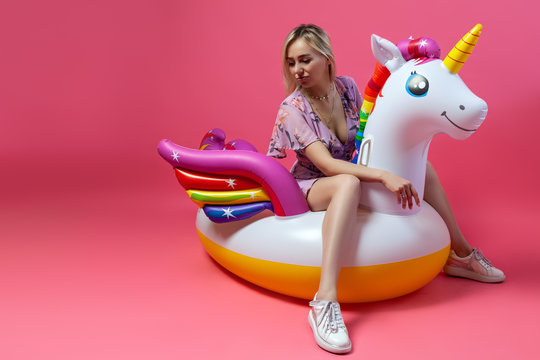 A blonde girl in a sexy sundress with slim legs in white sneakers sits on an inflatable multi-colored unicorn on a pink isolated background and look down. Summer vacation at the beach and travel.