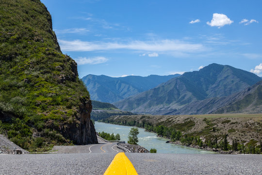 Close-up on a yellow dividing strip on an asphalt road chuysky tract stretching into the Altai Mountains near a rock with rocks and a river with a picturesque landscape in the background