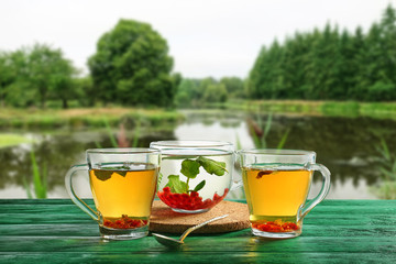 Cups of hot tea with berries on table near beautiful pond