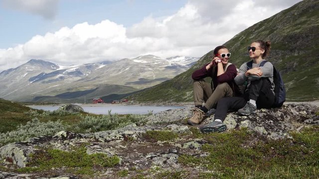 Two young women travelers in trekking boots resting sitting on the rock and enjoying a view of the Norwegian Fjord. Backpacker trip to Norway.