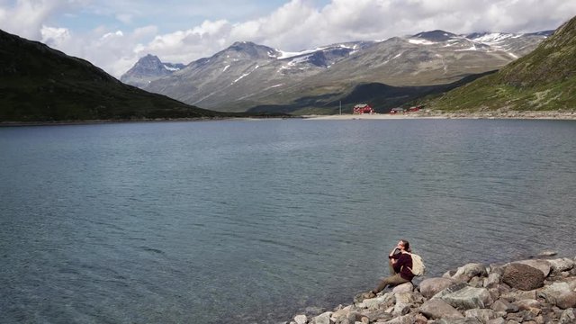 Young woman traveler resting sitting on a rock and enjoying a view of the mountain lake. Backpacker trip to Norway. Jotunheimen National Park.