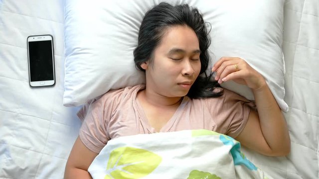 Top view of angry Asian young woman who had to wake up to answer the phone while sleeping on bed. Health care concept.