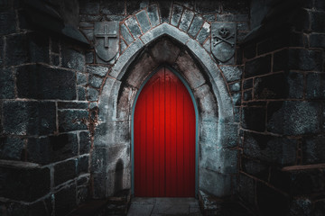 Scary pointy red wooden door in an old and wet stone wall building with cross, skull and bones at...