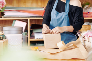 Cropped image of flower shop worker in blue apron wrapping gift box in decorative paper