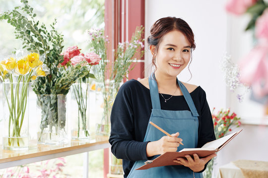 Portrait of pretty smiling young florist writing in planner and looking at camera