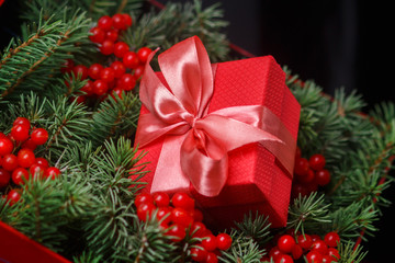 New Year Christmas Xmas 2020 holiday celebration red present gift box with satin pink bow, immersed in the needles of a Christmas tree decorated with red berries. Dark festive composition.